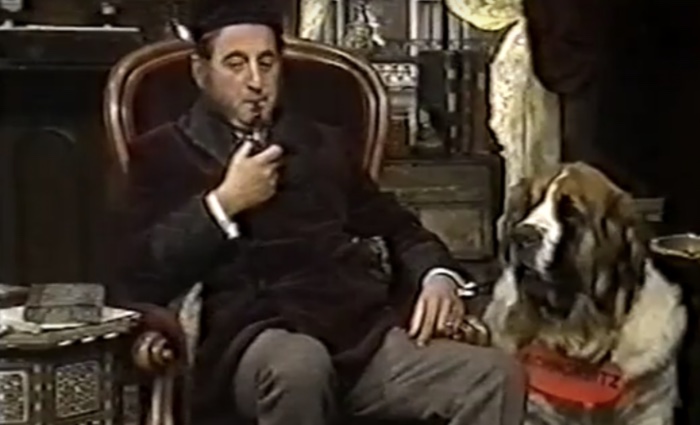 Bernie Winters in an armchair dressed as Sherlock Holmes, smoking a Meerschaum pipe, with his St. Bernard dog Schnorbitz sitting in front of him.